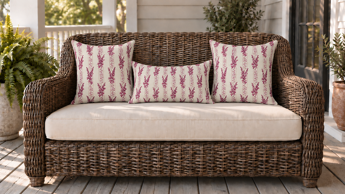 Two square outdoor pillows and a lumbar on a rattan loveseat on a wooden porch